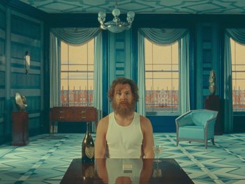 Movie still from “The Wonderful Story of Henry Sugar” (2023), directed by Wes Anderson – A man sitting at a table with a bottle of wine in front of him; Wide shot, Low angle