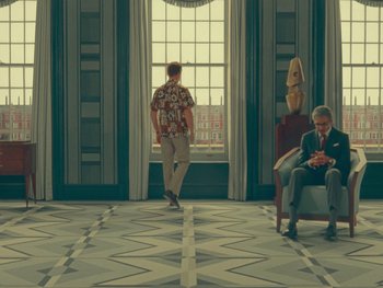 Movie still from “The Wonderful Story of Henry Sugar” (2023), directed by Wes Anderson – A man walking in front of a window while another man sits in a chair; Wide shot, Low angle