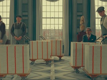 Movie still from “The Wonderful Story of Henry Sugar” (2023), directed by Wes Anderson – A painting of two men standing in front of boxes on dolly wheels; Wide shot, Over the shoulder angle