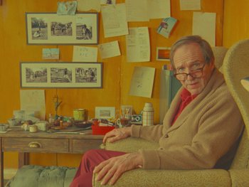 Movie still from “The Wonderful Story of Henry Sugar” (2023), directed by Wes Anderson – An older man sitting on a chair in a room with papers on the wall; Medium shot, Over the shoulder angle