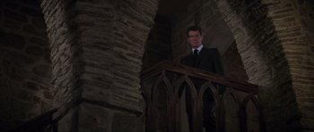 Movie still from “The World Is Not Enough” (1999), directed by Michael Apted – A man in a suit and tie standing on a railing in front of a stone wall; Medium shot, Low angle