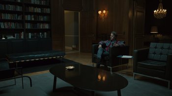 Movie still from “The Wrath of God” (2022), directed by Sebastián Schindel – A man sitting in a chair in a living room next to a coffee table; Wide shot, High angle