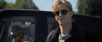 Movie still from “There's Someone Inside Your House” (2021), directed by Patrick Brice – A man with blonde hair wearing sunglasses sitting in the back of a car; Close Up shot, Over the shoulder angle