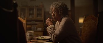 Movie still from “There's Someone Inside Your House” (2021), directed by Patrick Brice – An older woman sitting at a table with her hands clasped together; Close Up shot, Over the shoulder angle