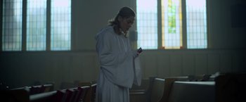 Movie still from “There's Someone Inside Your House” (2021), directed by Patrick Brice – A woman in a white robe is looking at her cell phone; Medium shot, Low angle