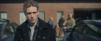Movie still from “There's Someone Inside Your House” (2021), directed by Patrick Brice – A man in a black jacket is standing in front of a brick building; Close Up shot, Over the shoulder angle