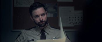 Movie still from “There's Someone Inside Your House” (2021), directed by Patrick Brice – A man in a police uniform looking at a paper; Close Up shot, Over the shoulder angle