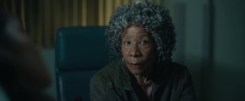 Movie still from “There's Someone Inside Your House” (2021), directed by Patrick Brice – An older woman sitting in front of a blue wall; Close Up shot, Over the shoulder angle