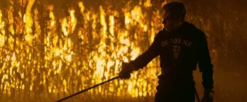 Movie still from “There's Someone Inside Your House” (2021), directed by Patrick Brice – A man holding a sword in front of a fire; Medium shot, Over the shoulder angle