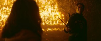 Movie still from “There's Someone Inside Your House” (2021), directed by Patrick Brice – A person holding a stick in front of a fire; Medium shot, Over the shoulder angle