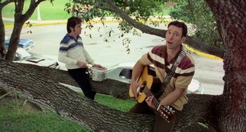 Movie still from “There's Something About Mary” (1998), directed by Bobby Farrelly – A man sitting on top of a tree playing a guitar; Medium shot, Low angle