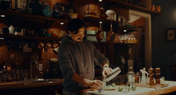 Movie still from “There's Something Wrong with the Children” (2023), directed by Roxanne Benjamin – A man is making a drink in a kitchen; Medium shot, Low angle