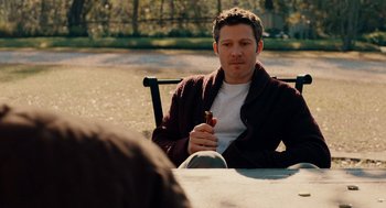 Movie still from “There's Something Wrong with the Children” (2023), directed by Roxanne Benjamin – A man sitting at a table holding a pipe in his hand; Medium shot, Over the shoulder angle