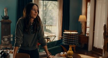Movie still from “There's Something Wrong with the Children” (2023), directed by Roxanne Benjamin – A woman standing at a table in a living room; Medium shot, Over the shoulder angle