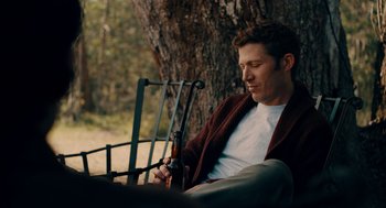 Movie still from “There's Something Wrong with the Children” (2023), directed by Roxanne Benjamin – A man sitting on a bench with a rifle in his hand; Medium shot, High angle