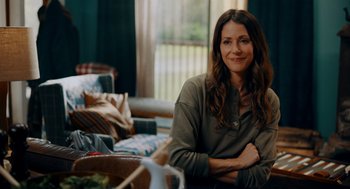 Movie still from “There's Something Wrong with the Children” (2023), directed by Roxanne Benjamin – A woman sitting on a couch in a living room; Medium shot, Over the shoulder angle