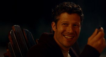 Movie still from “There's Something Wrong with the Children” (2023), directed by Roxanne Benjamin – A man smiling for the camera while sitting in a chair; Close Up shot, Over the shoulder angle