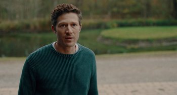 Movie still from “There's Something Wrong with the Children” (2023), directed by Roxanne Benjamin – A man in a green sweater standing in front of some grass; Medium shot, Over the shoulder angle