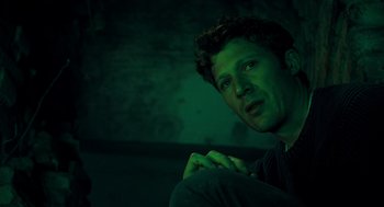 Movie still from “There's Something Wrong with the Children” (2023), directed by Roxanne Benjamin – A man sitting in front of a wall in a dark room; Close Up shot, Low angle