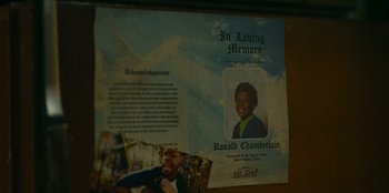 Movie still from “They Cloned Tyrone” (2023), directed by Juel Taylor – A funeral program with a picture of a young boy; Extreme Close Up shot, Overhead angle