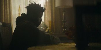 Movie still from “They Cloned Tyrone” (2023), directed by Juel Taylor – A man sitting at a table in a living room; Close Up shot, Over the shoulder angle