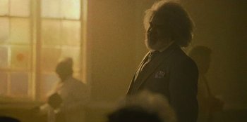 Movie still from “They Cloned Tyrone” (2023), directed by Juel Taylor – An old man with a beard is standing in a darkened room; Close Up shot, Over the shoulder angle