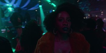 Movie still from “They Cloned Tyrone” (2023), directed by Juel Taylor – A woman in a brown jacket is standing in a dark room; Close Up shot, Over the shoulder angle