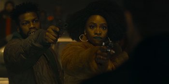 Movie still from “They Cloned Tyrone” (2023), directed by Juel Taylor – A woman holding two guns in her hands; Close Up shot, Over the shoulder angle
