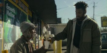 Movie still from “They Cloned Tyrone” (2023), directed by Juel Taylor – A man handing a drink to a homeless person; Medium shot, Low angle