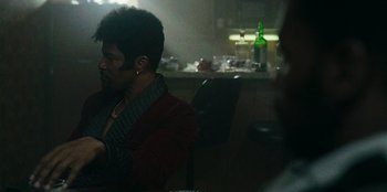 Movie still from “They Cloned Tyrone” (2023), directed by Juel Taylor – A man sitting at a table with a bottle in front of him; Medium shot, Over the shoulder angle