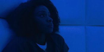 Movie still from “They Cloned Tyrone” (2023), directed by Juel Taylor – A young woman is sitting in a dark room; Close Up shot, Low angle