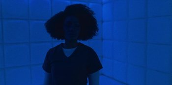 Movie still from “They Cloned Tyrone” (2023), directed by Juel Taylor – A woman standing in a room lit up by a blue light; Medium shot, Low angle