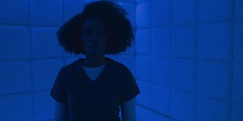 Movie still from “They Cloned Tyrone” (2023), directed by Juel Taylor – A woman with a very large afro stands in a room; Medium shot, Low angle