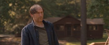 Movie still from “They/Them” (2022), directed by John Logan – A man standing in front of a house in the woods; Medium shot, Over the shoulder angle