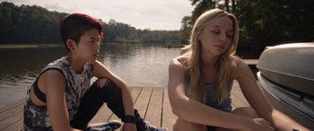 Movie still from “They/Them” (2022), directed by John Logan – Two young women sitting on a dock near a body of water; Medium shot, Over the shoulder angle