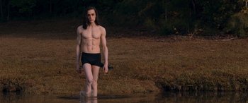 Movie still from “They/Them” (2022), directed by John Logan – A man with long black hair is standing in the water; Medium shot, Low angle