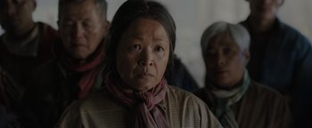 Movie still from “Thirteen Lives” (2022), directed by Ron Howard – An older woman wearing a neck scarf looking at the camera; Close Up shot, Over the shoulder angle