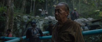 Movie still from “Thirteen Lives” (2022), directed by Ron Howard – An old man standing in the woods with people in the background; Close Up shot, Over the shoulder angle