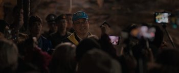 Movie still from “Thirteen Lives” (2022), directed by Ron Howard – A man wearing a blue hat is taking a picture of a crowd; Medium shot, Over the shoulder angle