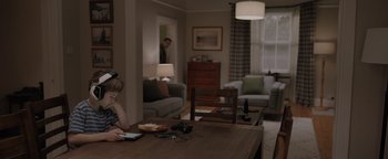 Movie still from “Thirteen Lives” (2022), directed by Ron Howard – A woman sitting at a wooden table in a living room; Wide shot, Over the shoulder angle