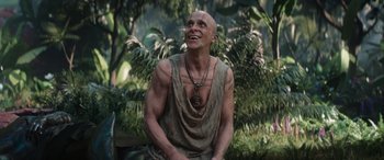 Movie still from “Thor: Love and Thunder” (2022), directed by Taika Waititi – A man with a shaved head sitting on top of a wooden bench; Medium shot, Low angle