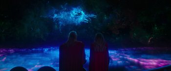 Movie still from “Thor: Love and Thunder” (2022), directed by Taika Waititi – Two women in red robes looking at a projection of a dragon; Extreme Wide shot, Overhead angle