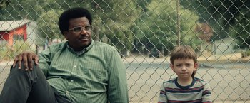 Movie still from “Timmy Failure: Mistakes Were Made” (2020), directed by Tom McCarthy – A man and a boy sitting next to each other in front of a chain link fence; Close Up shot, Over the shoulder angle