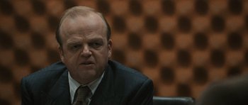 Movie still from “Tinker Tailor Soldier Spy” (2011), directed by Tomas Alfredson – A man in a suit sitting in front of an audience; Close Up shot, Over the shoulder angle