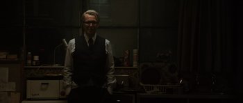 Movie still from “Tinker Tailor Soldier Spy” (2011), directed by Tomas Alfredson – A man standing in a dark room wearing a vest; Medium shot, Over the shoulder angle