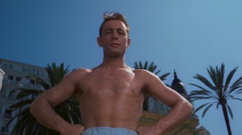 Movie still from “To Catch a Thief” (1955), directed by Alfred Hitchcock – A shirtless man standing with his hands on his hips in front of palm trees; Medium shot, Low angle