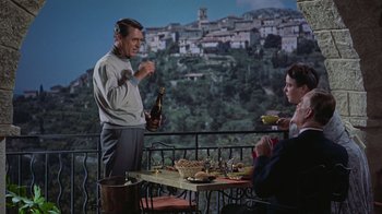 Movie still from “To Catch a Thief” (1955), directed by Alfred Hitchcock – A man standing next to a table holding a wine bottle; Medium shot, High angle