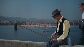 Movie still from “To Catch a Thief” (1955), directed by Alfred Hitchcock – An older man sitting on a wall holding a fishing pole; Medium shot, High angle