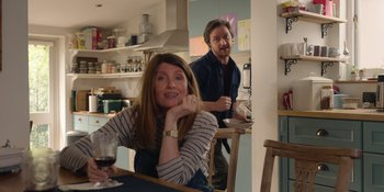Movie still from “Together” (2021), directed by Justin Martin – A woman sitting at a table with a man standing behind her; Medium shot, Over the shoulder angle