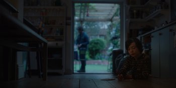 Movie still from “Together” (2021), directed by Justin Martin – A young boy sitting at a table in front of an open patio door; Wide shot, Over the shoulder angle